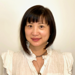 Headshot of woman with shoulder length dark hair, wearing a white short-sleeve blouse, standing against a beige background